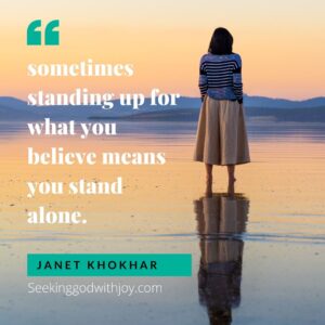 A quote by Janet Khokhar set on a photo of a woman standing on a beach at late sunset. The quote says, "sometimes standing up for what you believe means you stand alone."